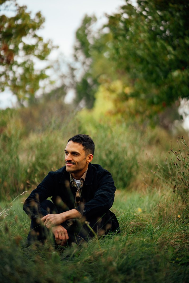 A man with dark hair and facial hair wearing a black shirt sitting in a field of grass and trees 
