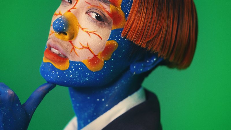 A person with mostly blue facepaint with a mask of a light shade around their face with different patterns of red and blue, and an orange wig, in front of a green screen