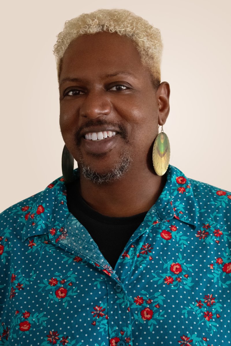 A person with dark skin and short blonde hair, earrings and a blue shirt smiling at the camera
