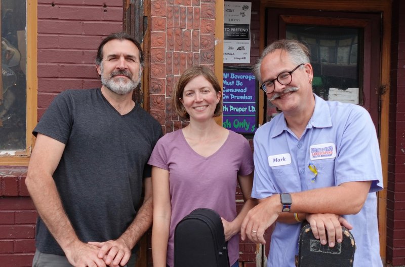Two older men and one woman with light skin tones and short sleeve shirts smile in front of a brick wall