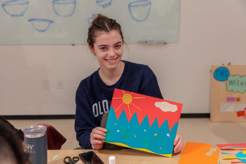 A teenaged girl with dark hair and light skin wearing a long sleeve black shirt holds up an art project