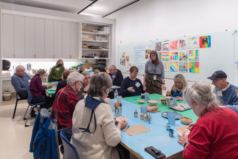A large group of elderly people in a studio painting ceramics while an instructor stands and speaks