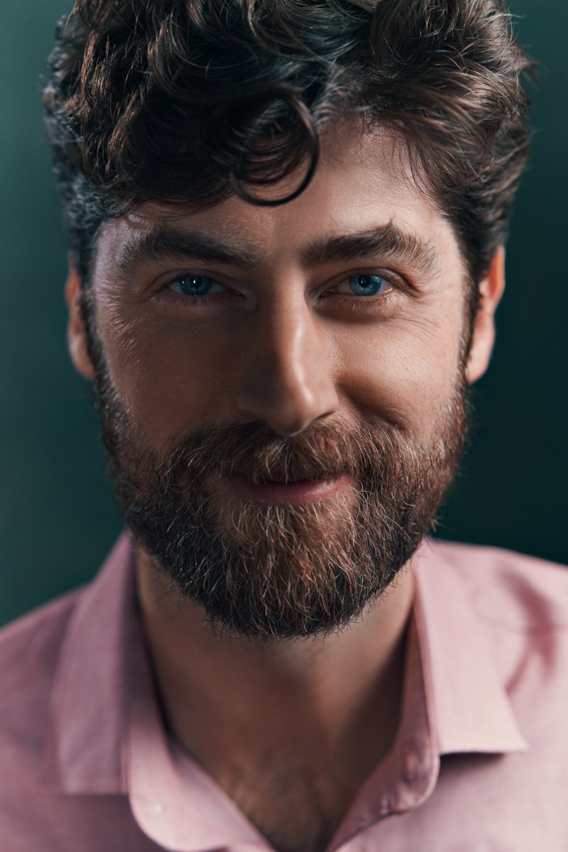 Closeup photo of a man in a pink shirt, with fair skin, green eyes, brown hair and beard, in front of a green backdrop