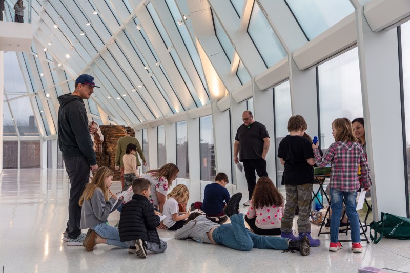 A group of children sitting on the ground drawing with a few adults around them in a large white hallway with big windows