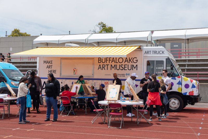 A white truck with a yellow awning and groups of people sitting at a table and around the truck making art projects