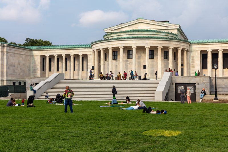 A museum building with marble columns and people walking around laying in the lawn out front
