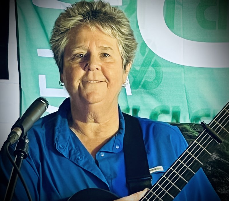 A woman with a blue shirt, light short hair, light skin tone, smiling while holding a guitar