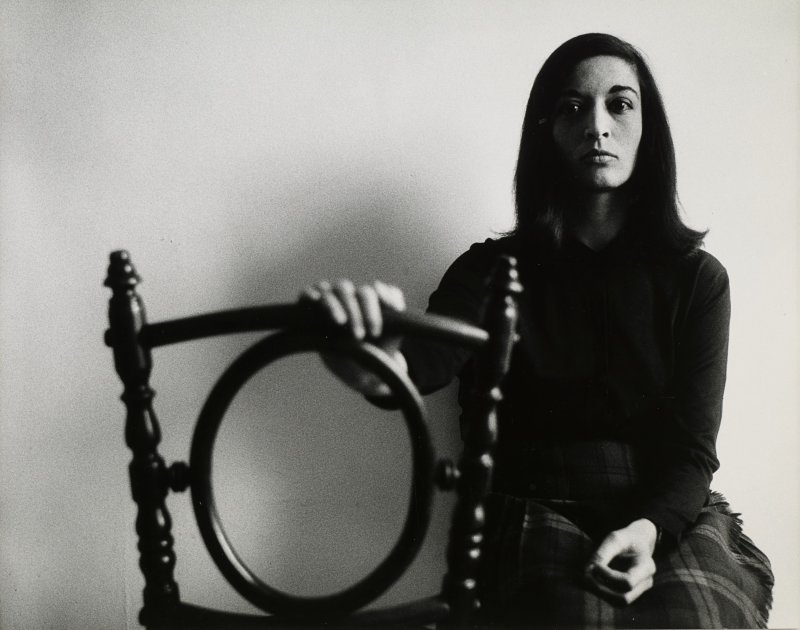 A black and white photo of a woman with dark hair and. black sweater and pants sitting with her hand resting on the back of a wooden chair
