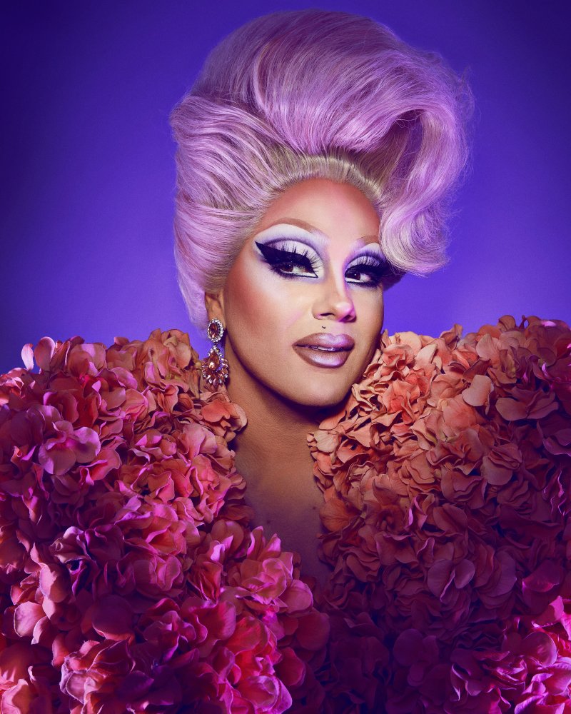 Headshot of a drag queen with a purple updo, bright eyemakeup, and a feather pink and orange top
