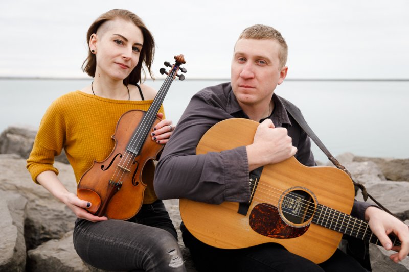 A woman with short brown hair that is shaved on one side, light skin tone, and holding a violin sits next to a man with short blonde hair, light skin tone, and is holding a guitar