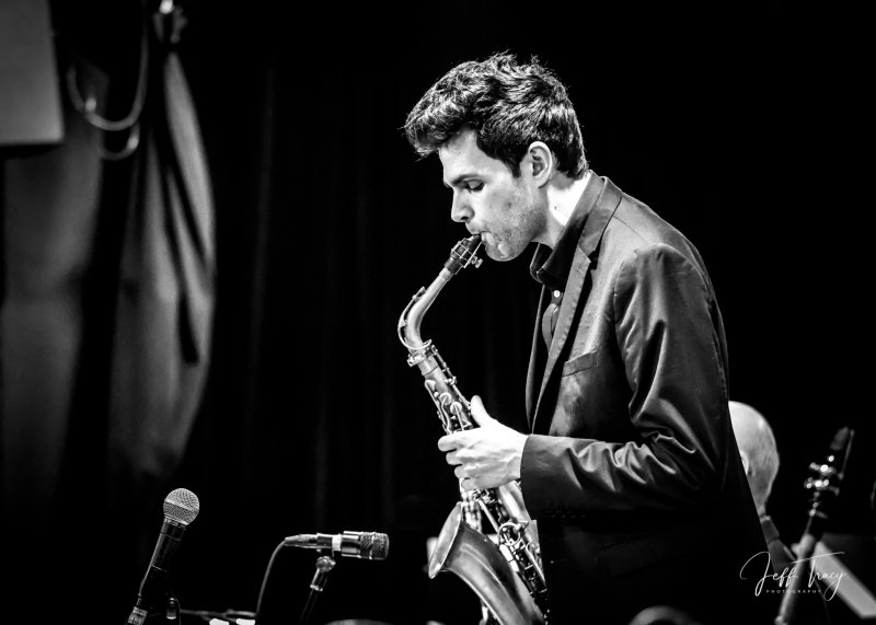 Black and white photo of a man with brown hair, light skin tone, wearing a black shirt, playing the saxophone