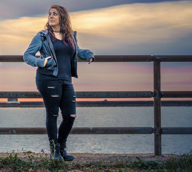A woman with brown hair, light skin tone, wearing a black shirt and ripped jeans, and a jean jacket, standing next to a railing in front of a sunset