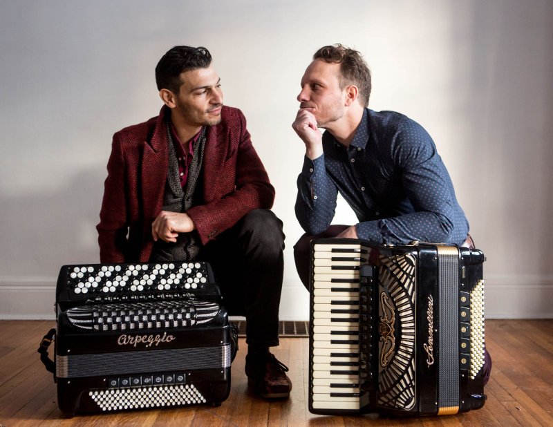 Two men sitting in front of piano accordians looking at one another