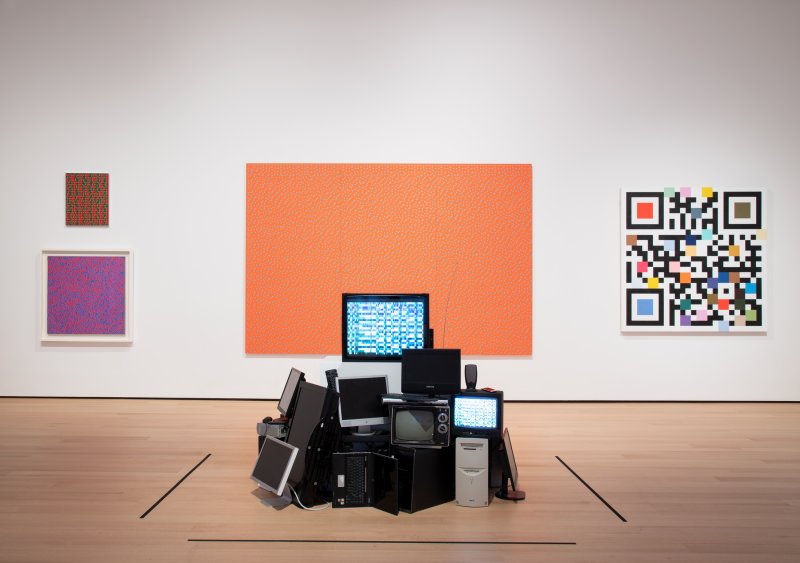 Gallery installation view of Op art paintings on a wall foregrounded by a pile of computers and tech equipment