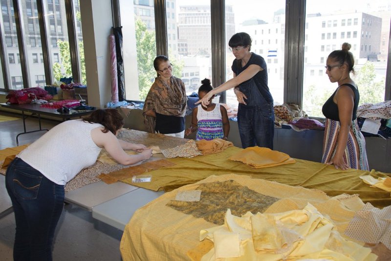 A group of people in a workshop looking at a large piece of yellow fabric draped on a table