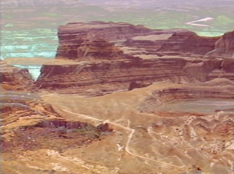 A dramatic view from above of a desert landscape, featuring sedimented rock formations and perhaps a lake.