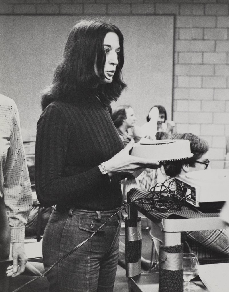 Black and white image of a person with long dark hair wearing a dark sweater and jeans in a classroom preparing a slide lecture