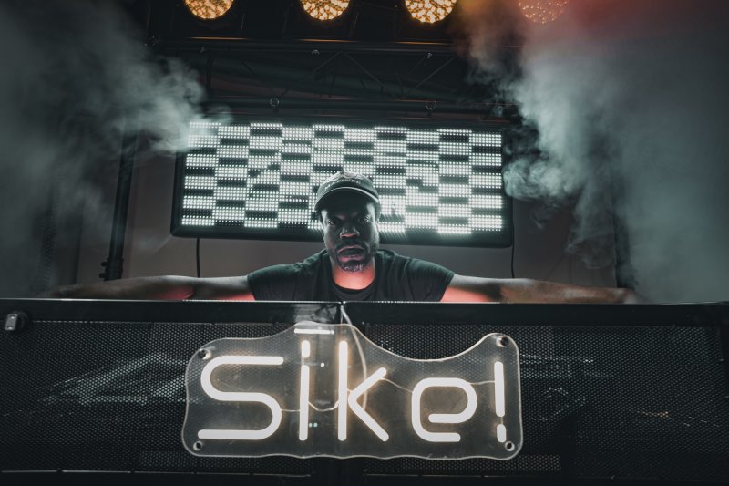 Dark image with vibrant lights of a person leaning over a DJ booth with the word "Sike!" illuminated in the front