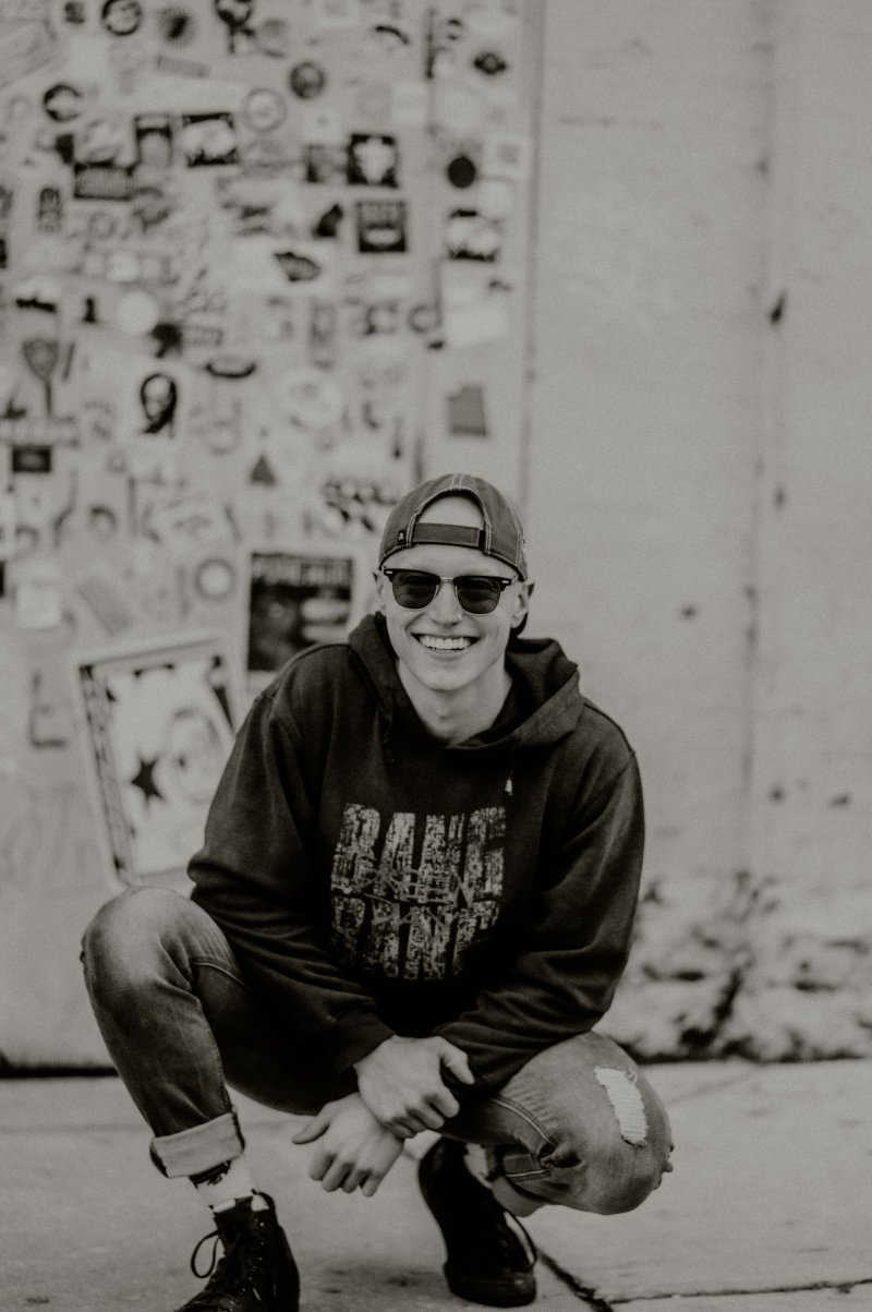 Black and white image of a person crouched down wearing black sunglasses and a backwards cap