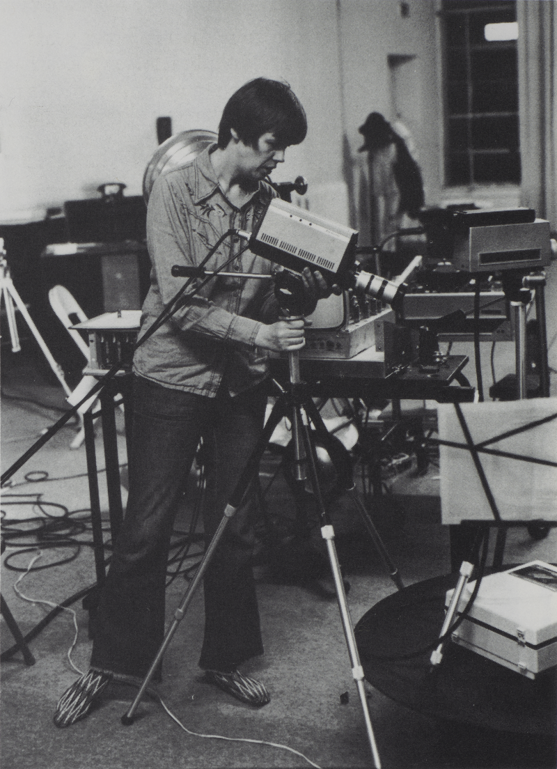 A person in a room full of technical equipment stands next a video camera mounted on a tripod