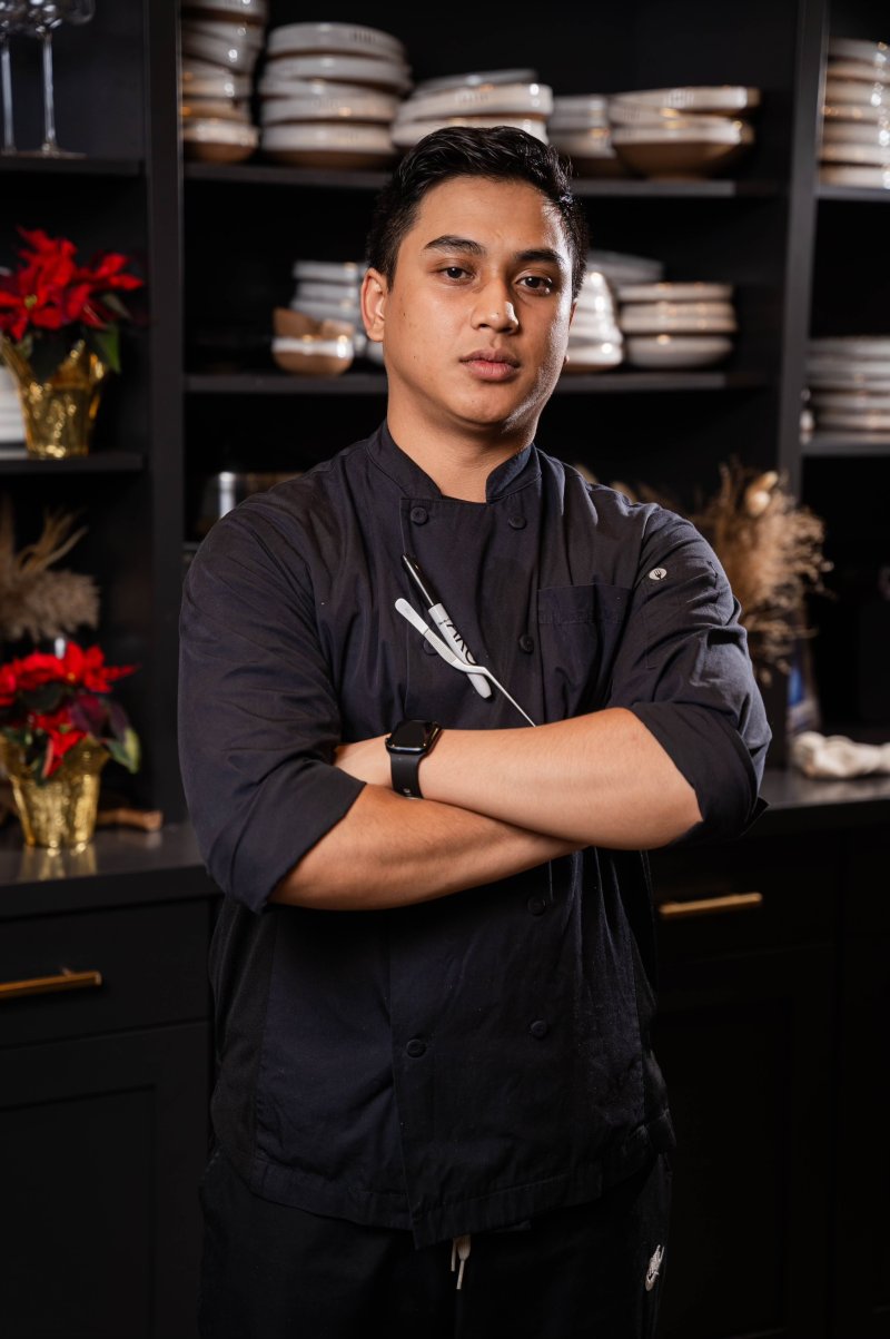 A chef posing in front of a kitchen in a black uniform with their arms crossed