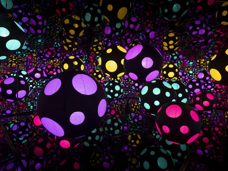 A Mirror Room installation that is dark with various black circles with colorful polka dots on them throughout 