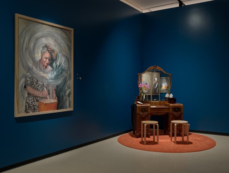 Installation view of a gallery with dark blue walls, an oil painting portrait, and an antique vanity in the corner of the space