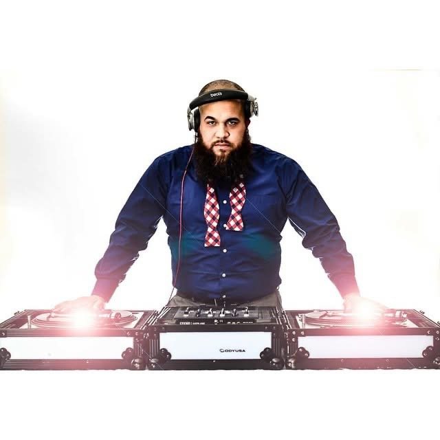 A man with a beard standing in front of two DJ turntables