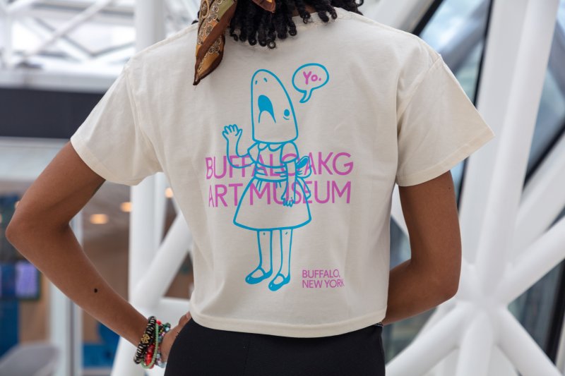 The back of a white shirt that has a blue shark girl illustration and "Buffalo AKG Art Museum" in pink font over top