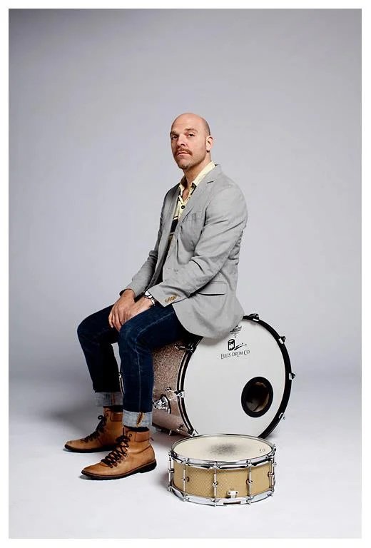 A person sitting on a drum, wearing a suit, in front of a grey background