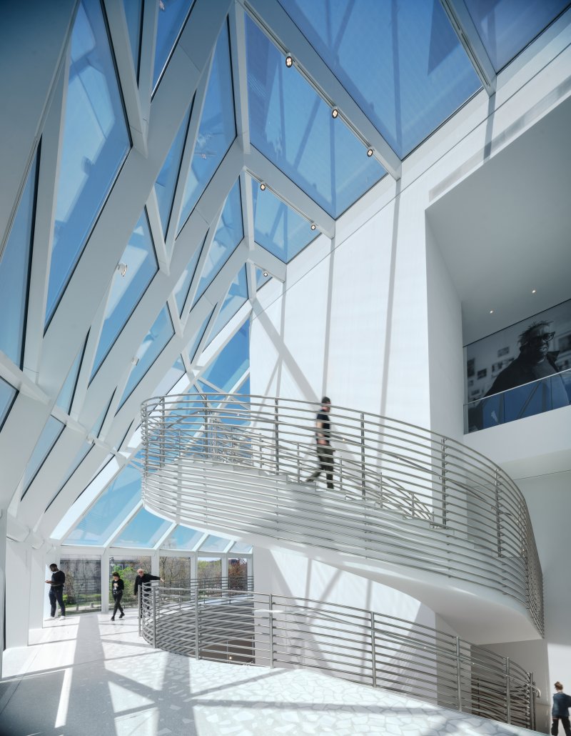 A winding staircase descending down a white wall with a glass ceiling overhead