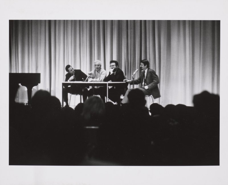 Black and white image of four people speaking on a stage