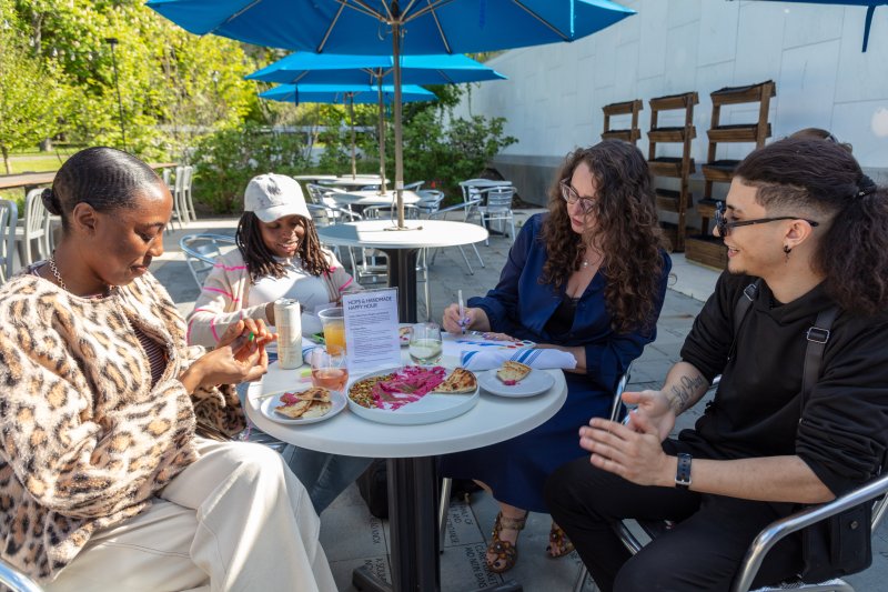 Four people enjoying drinks and crafts on the patio under a blue umbrella