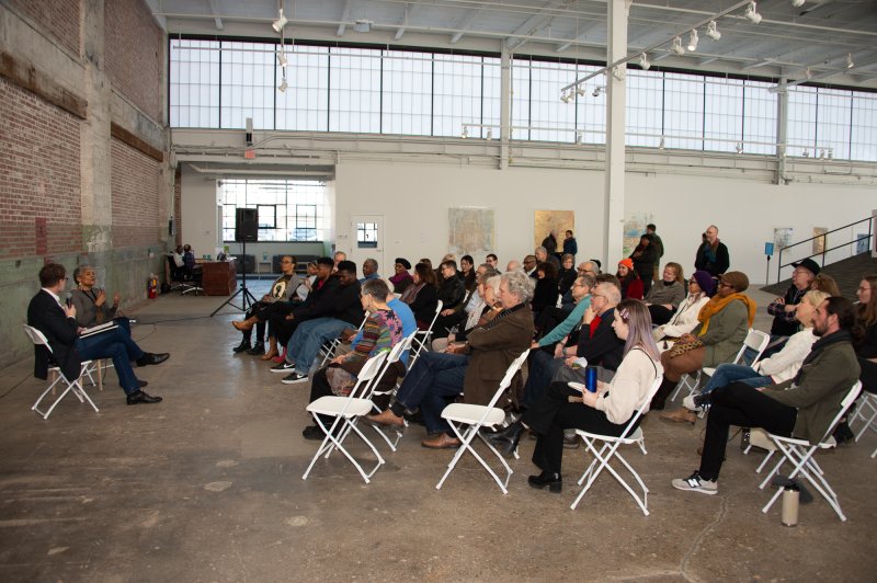 An African American woman and a white man talk in front of a group of people