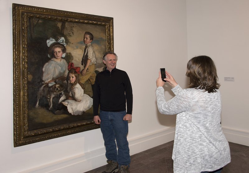 A woman in a white sweater taking a photo of a man in a dark sweater and jeans posing in front of a painting featuring three children from the early 1900s