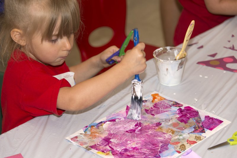 A young girl in a red shirt painting glue over a colorful pink collage