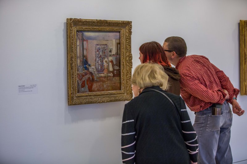 Two women and a man lean in to look at a square painting of a man and a child in a lavishly decorated room
