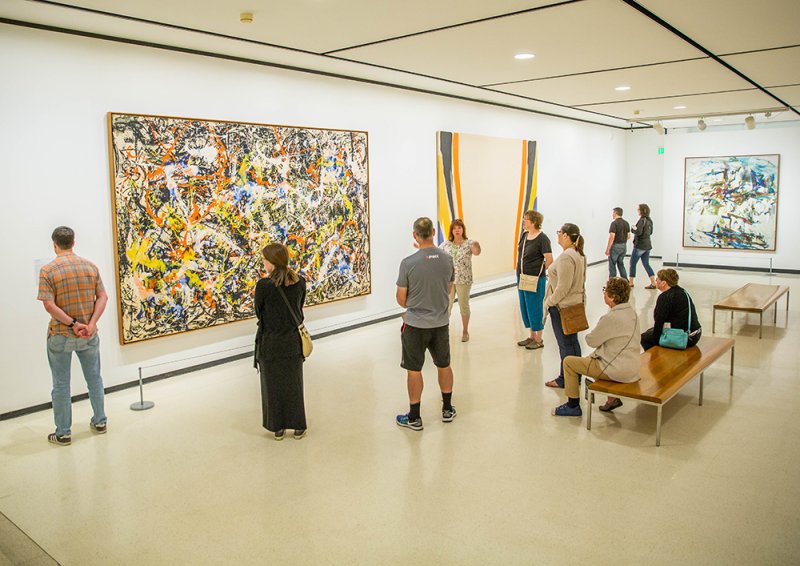 Guests on a tour in front of a large drip painting by Jackson Pollock featuring oranges, blues, yellows, and black