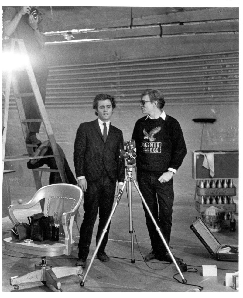 A black-and-white photograph of Gerard Malanga standing next to Andy Warhol