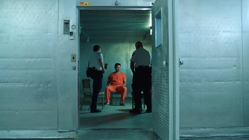 A man in an orange jumpsuit seated in a chair flanked by two prison guards