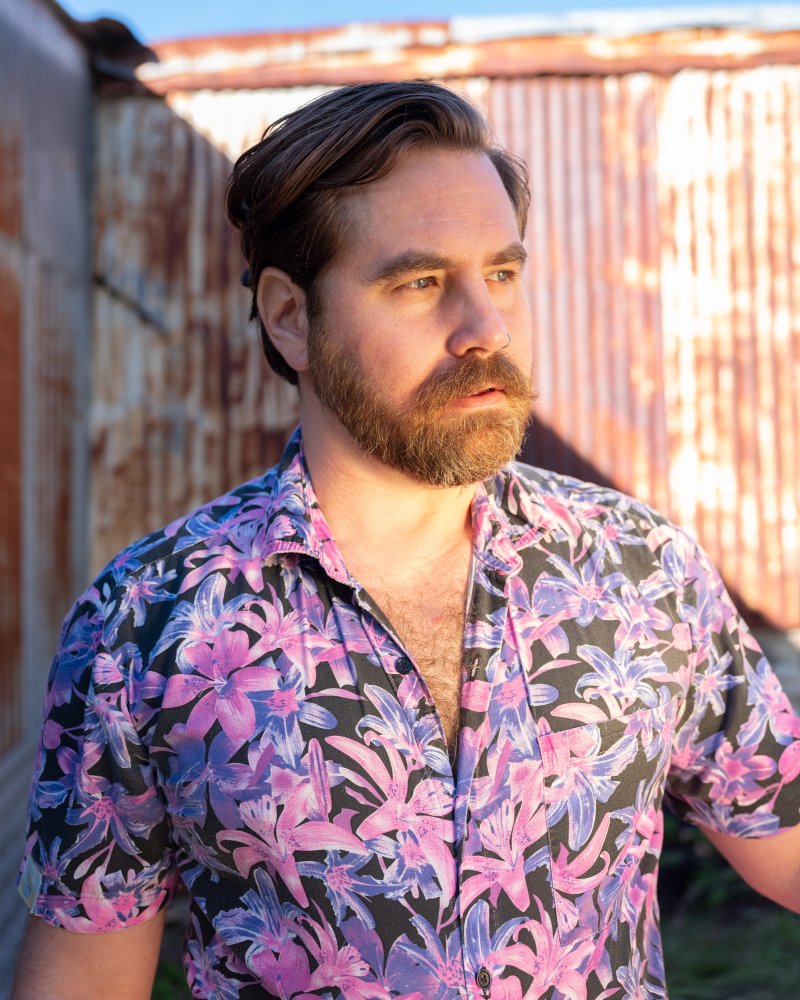 Photograph of a man with brown hair, a beard, and mustache, wearing a button down shirt with pink and purple flowers on a black background