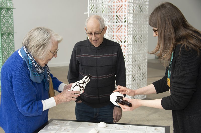 An older woman and a younger woman hold glass bottles with clay seashells glued to them while an older man looks on