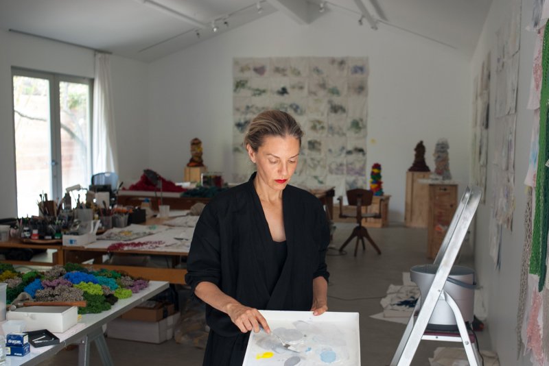 A woman with dirty blonde hair and a black shirt stands in a large room with artmaking materials holding a palette with paint on it and a palette knife