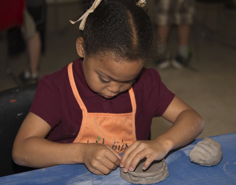 A young girl in an orange apron molding clay into a bowl using coils of clay