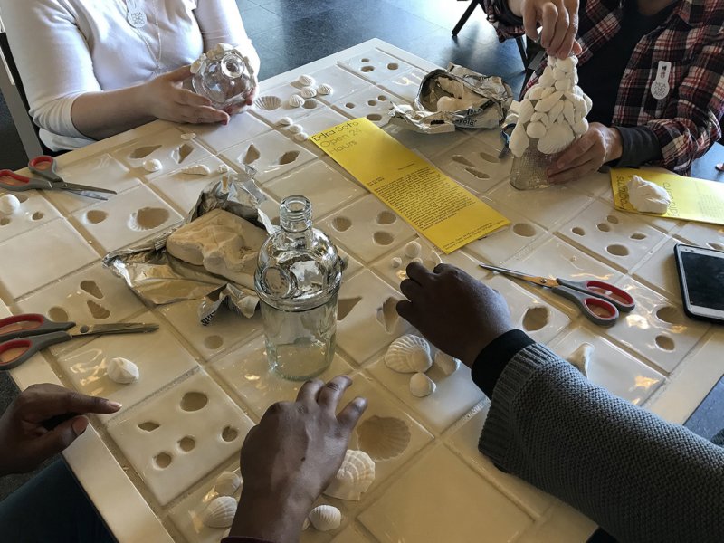 Four people affixing white cast shells to glass liquor bottles around a table