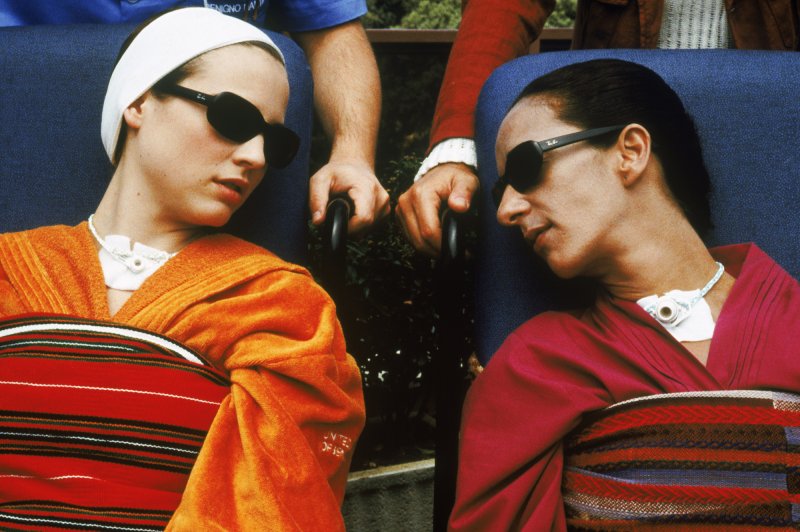 Two women wearing sunglasses, seated and talking to each other