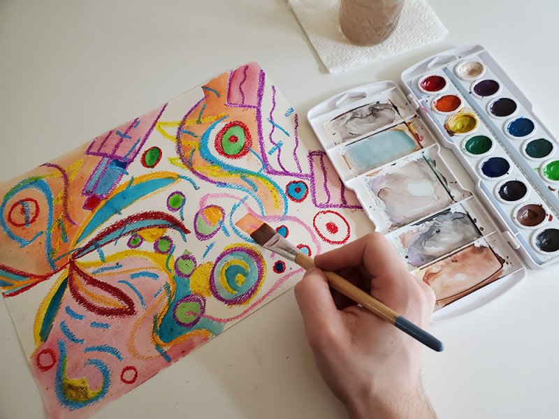 A person painting with watercolors on an oil pastel drawing