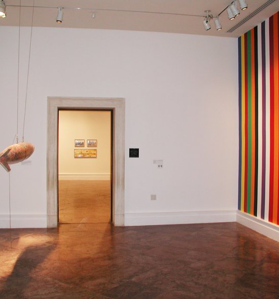 Installation view of a gallery with a rainbow striped wall and a hanging sculpture