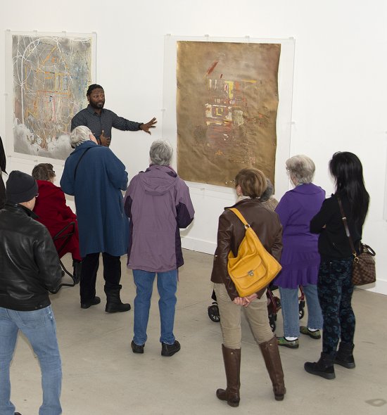 An African American man talks to a group of adults in front of a gold-colored painting