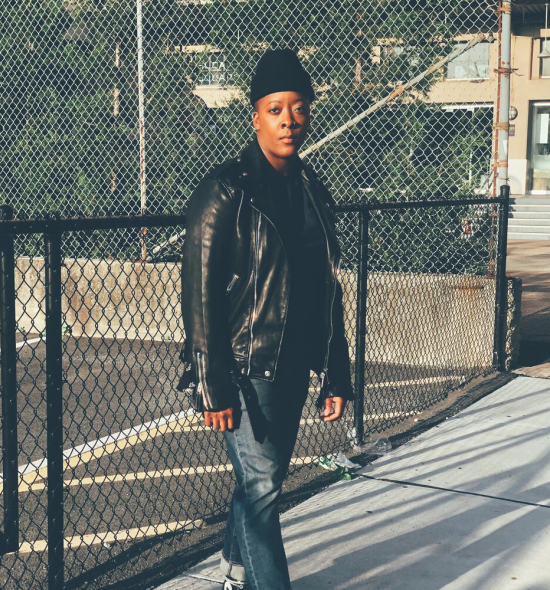 Photo of dark-skinned woman in leather jacket and black hat blacktop and fence in the background